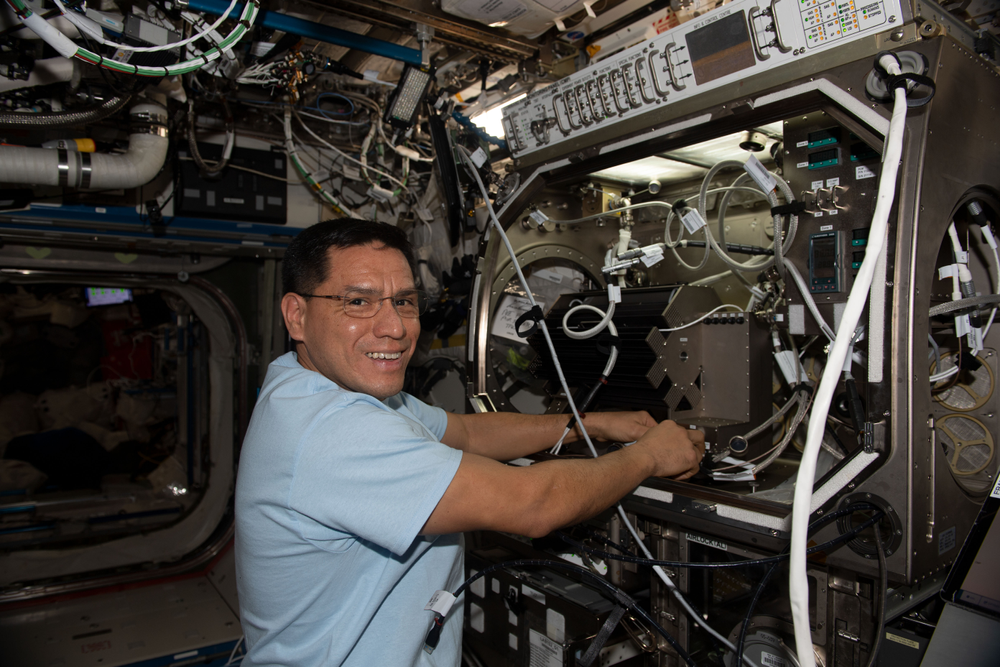 A male astronaut wearing glasses works inside a metal experiment box.