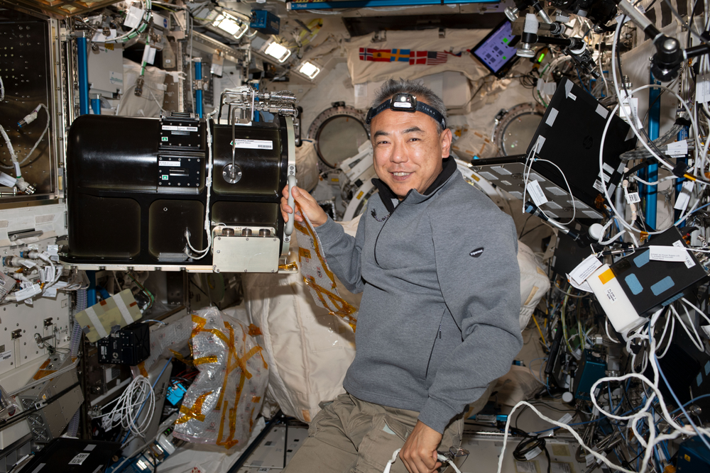 View of a person in microgravity wearing a grey jacket and a headlamp surrounded by wire and technology aboard the International Space Station.