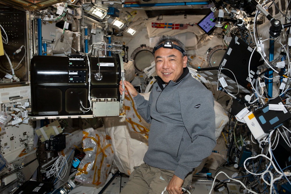 Astronaut smiles for the camera while floating inside a module on the International Space Station, surrounded by cables, instruments, and equipment. 