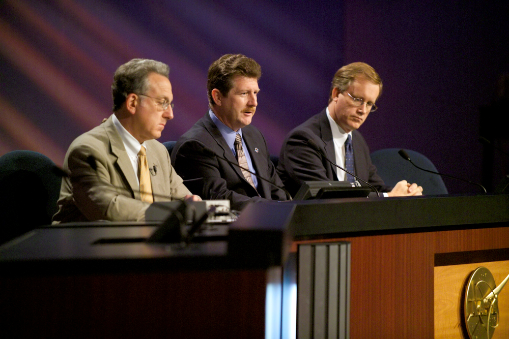 Three people are sitting at a desk with a personal microphone near their face. The background is a gradient of purple.  