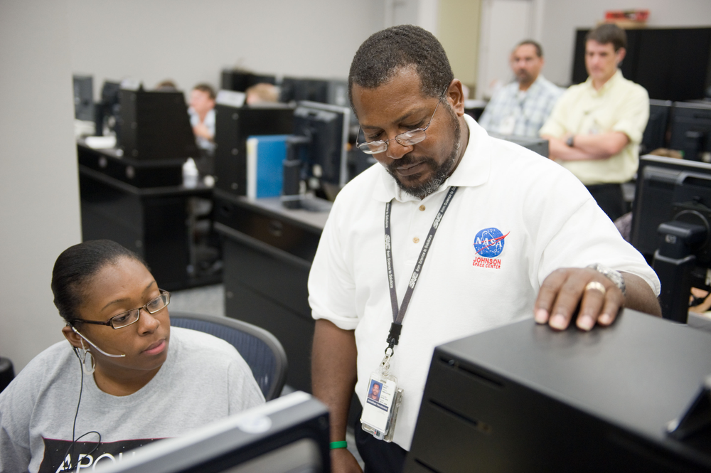 Two people in front of a desk looking at a monitor. 