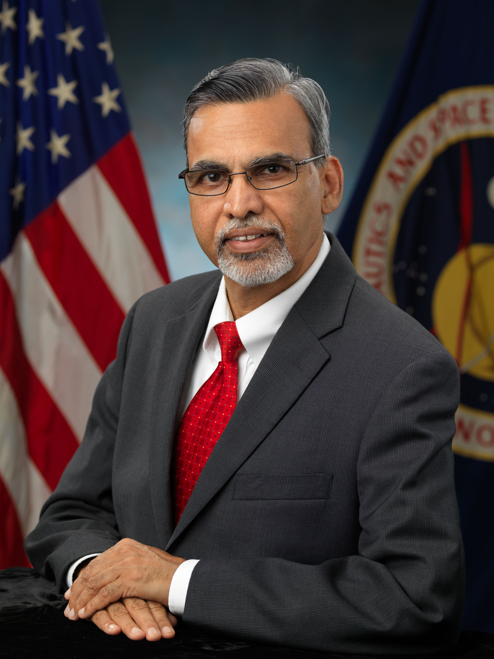 A person wearing a grey suit, white button-down, and red tie in front of a blue/grey background with two flags behind him. 