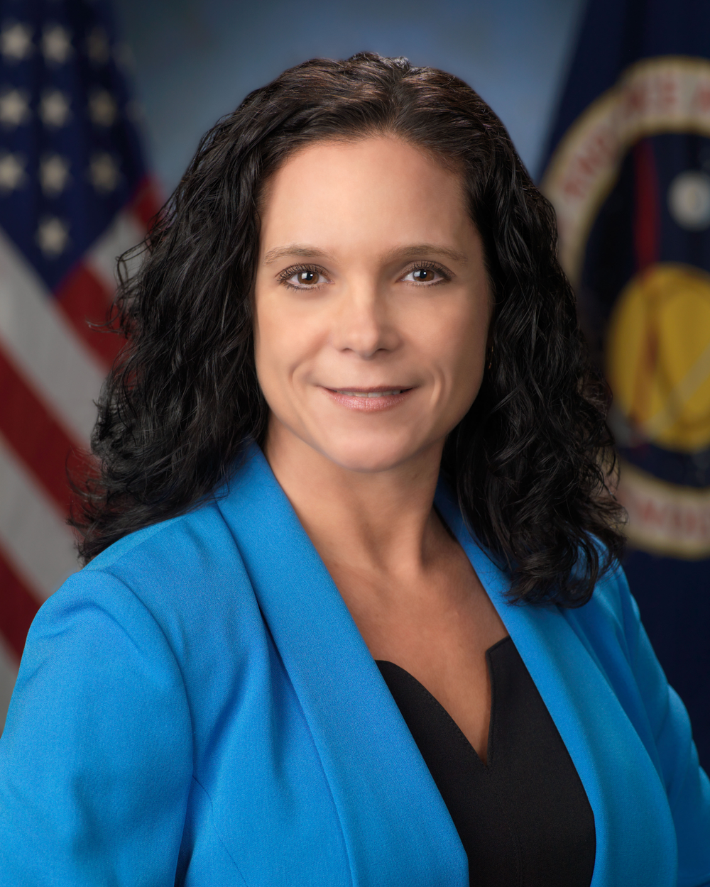 A portrait of a woman with long, dark, wavy hair, wearing a bright blue blazer over a black top. The background includes the American flag and a NASA emblem.