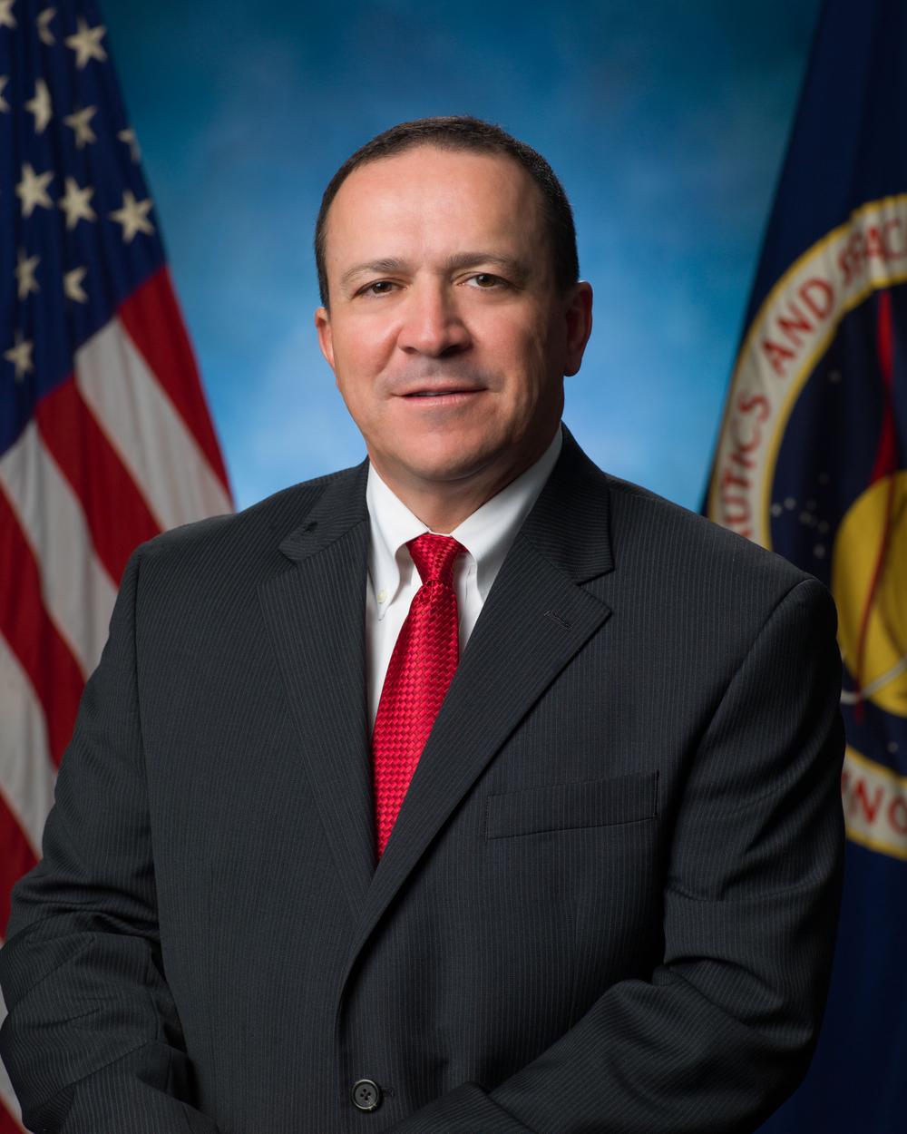 A person wearing a black suit and red tie smiles in front of a blue background and a U.S. flag (left) and NASA flag (right).