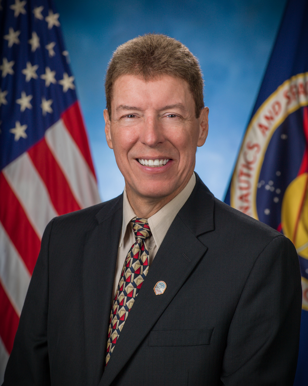 A person wearing a black suit and multicolored tie smiles in front of a blue background and a U.S. flag (left) and NASA flag (right).