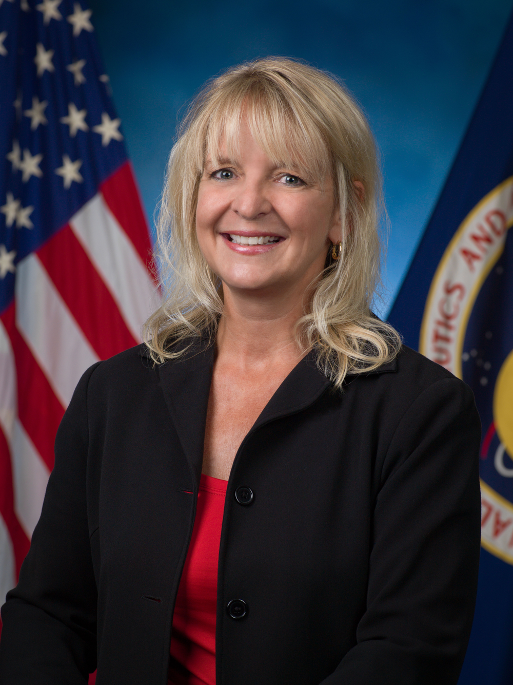 A person wearing a black suit and red blouse smiles in front of a blue background and a U.S. flag (left) and NASA flag (right).