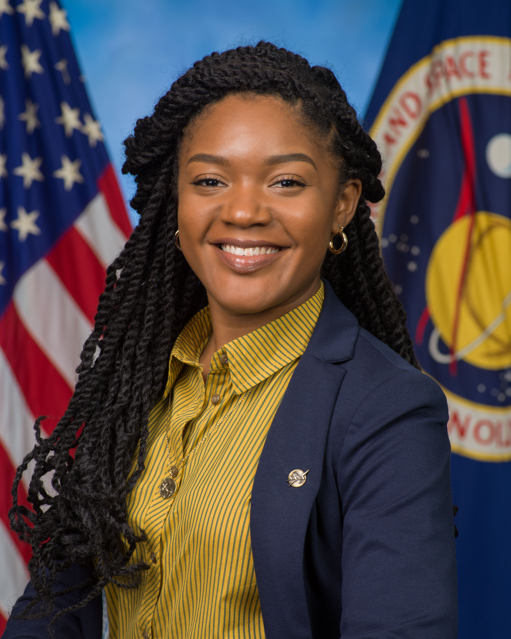 An image of a person in a yellow striped button down and black blazer with a NASA and American flag in the background. 