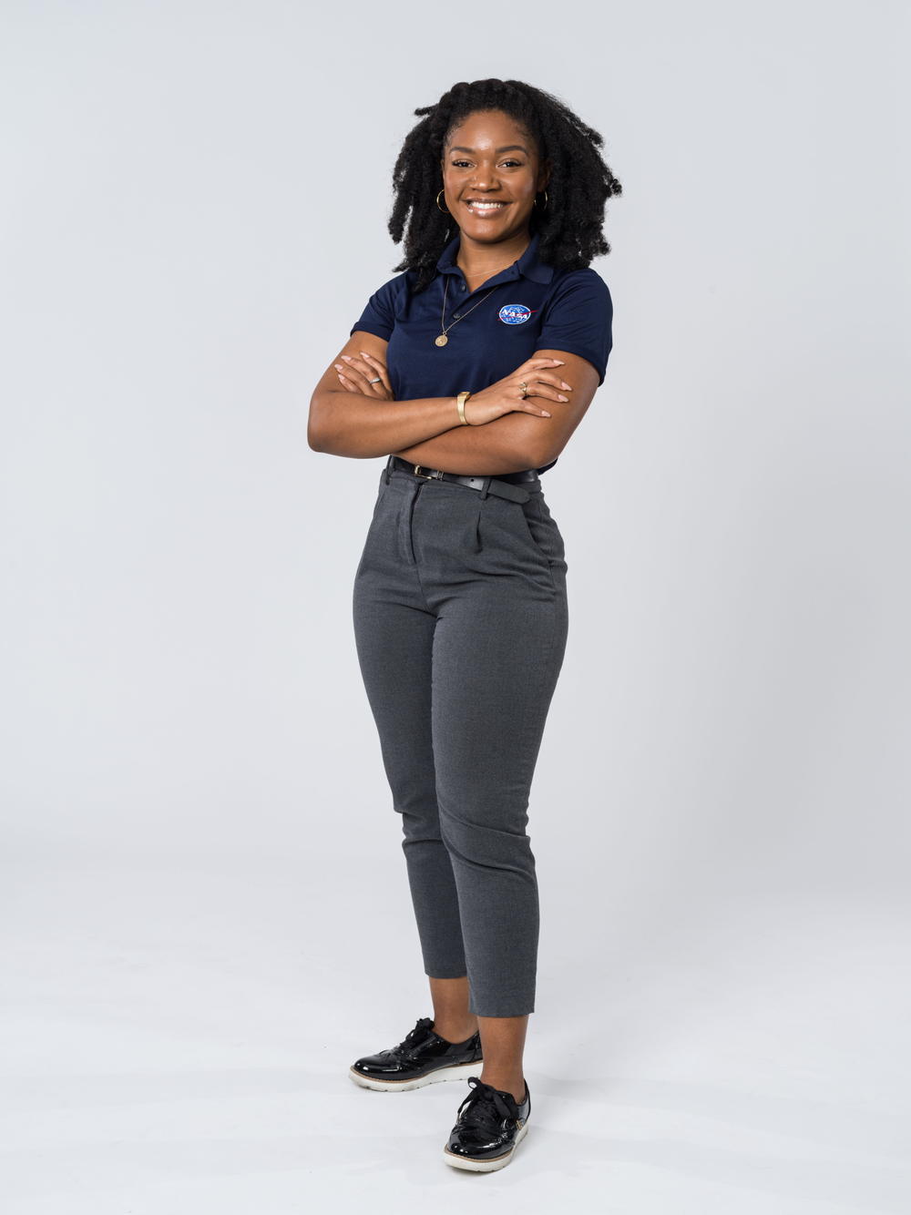 Image of a person in a blue shirt with a NASA meatball emblem and grey pants smiling in front of a grey background. 