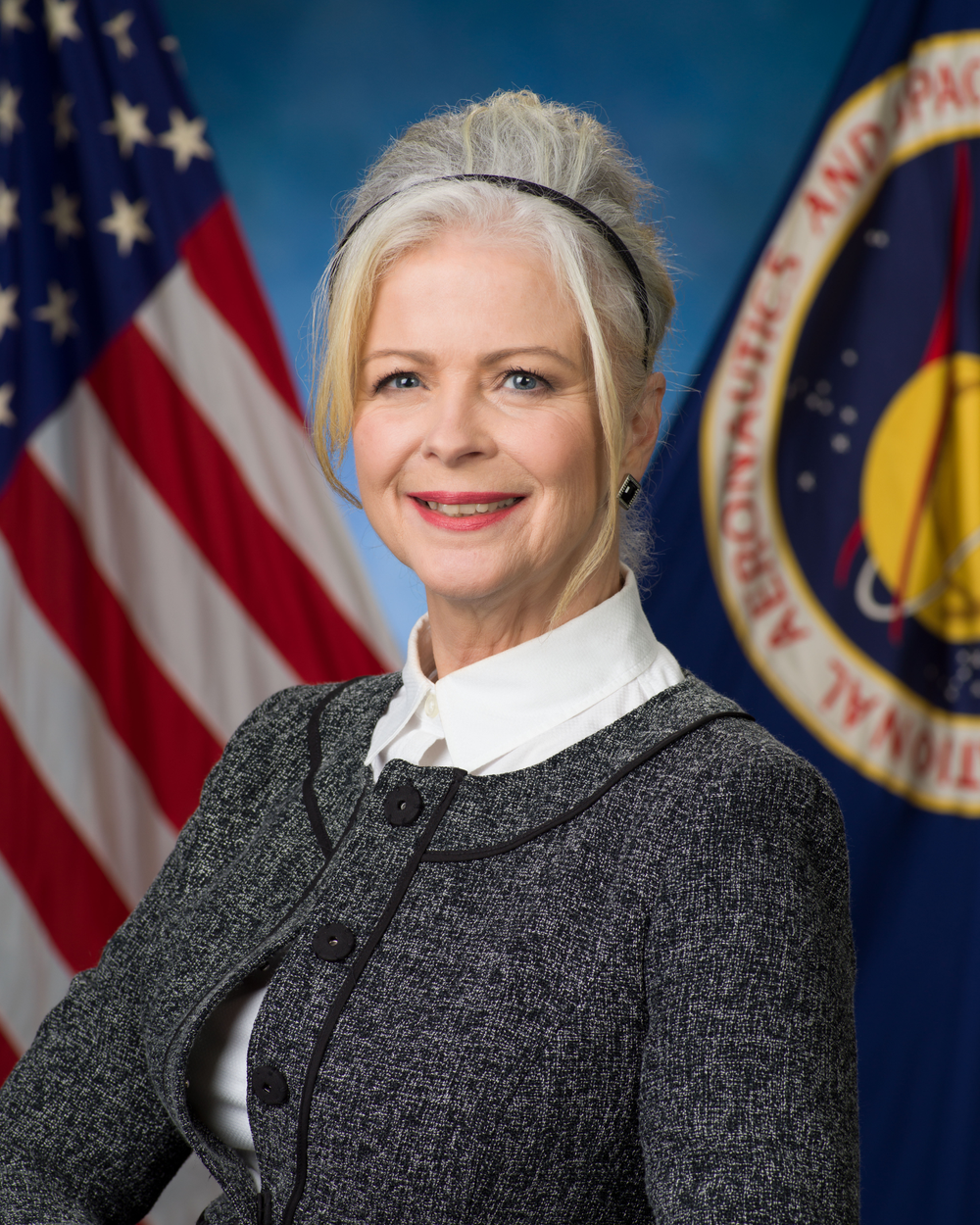 A professional headshot of a woman with white hair with an American flag in the background.