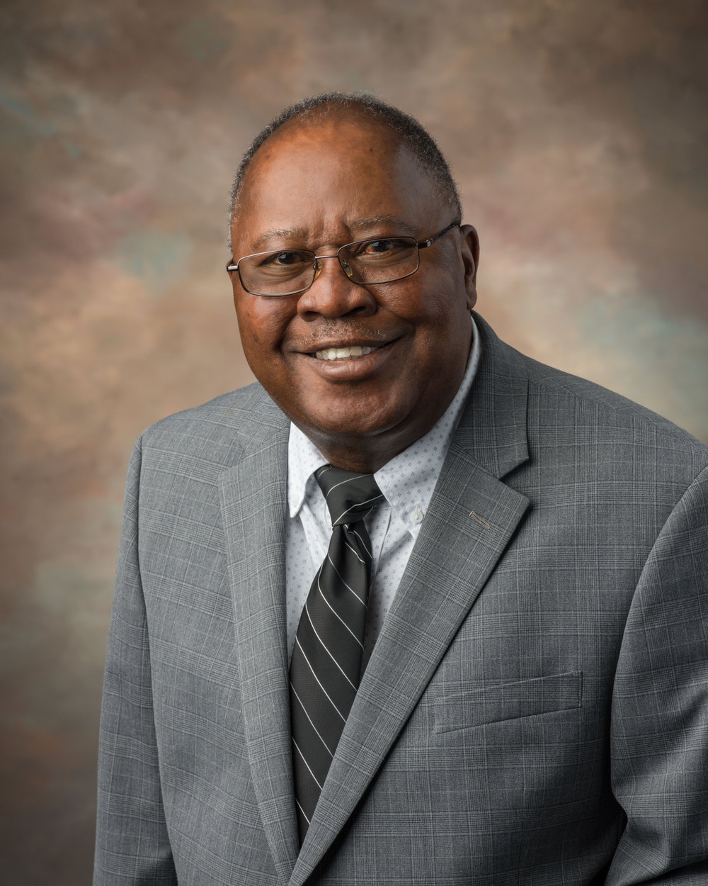 An official portrait of a Black man wearing glasses, a light gray suit, and a dark gray striped tie.