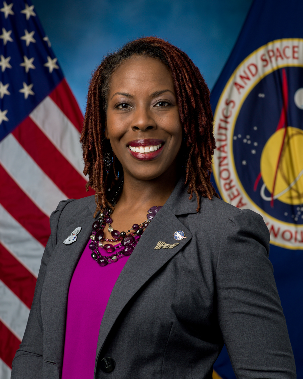 A woman wearing a grey blazer and a pink shirt smiles in front of a blue background with two flags behind her,  a U.S. flag on the left and a NASA flag on the right.