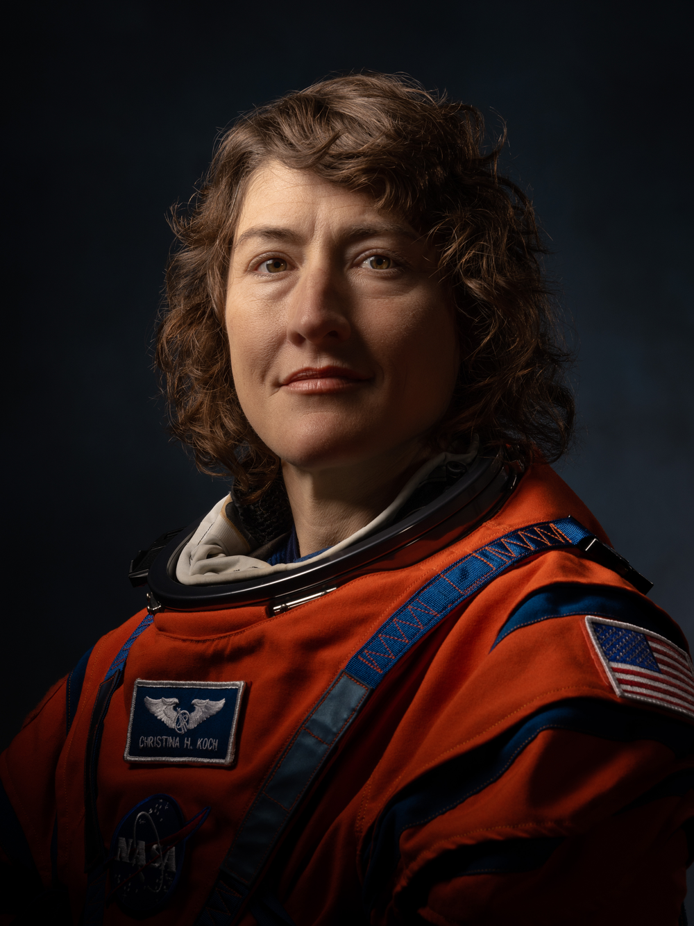 A portrait of a female astronaut with short, curly brown hair, wearing an orange NASA spacesuit in front of a black background. The suit features an American flag patch on the shoulder and NASA insignia on the chest. 