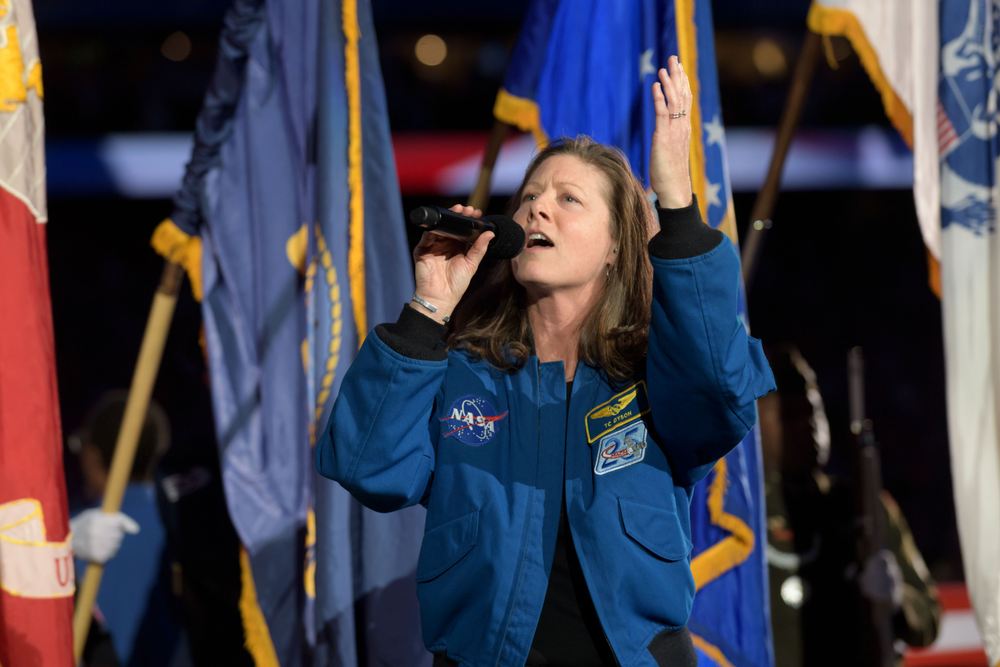 A female astronaut wearing a blue astronaut coat holds a microphone and sings with flags waving in the background.