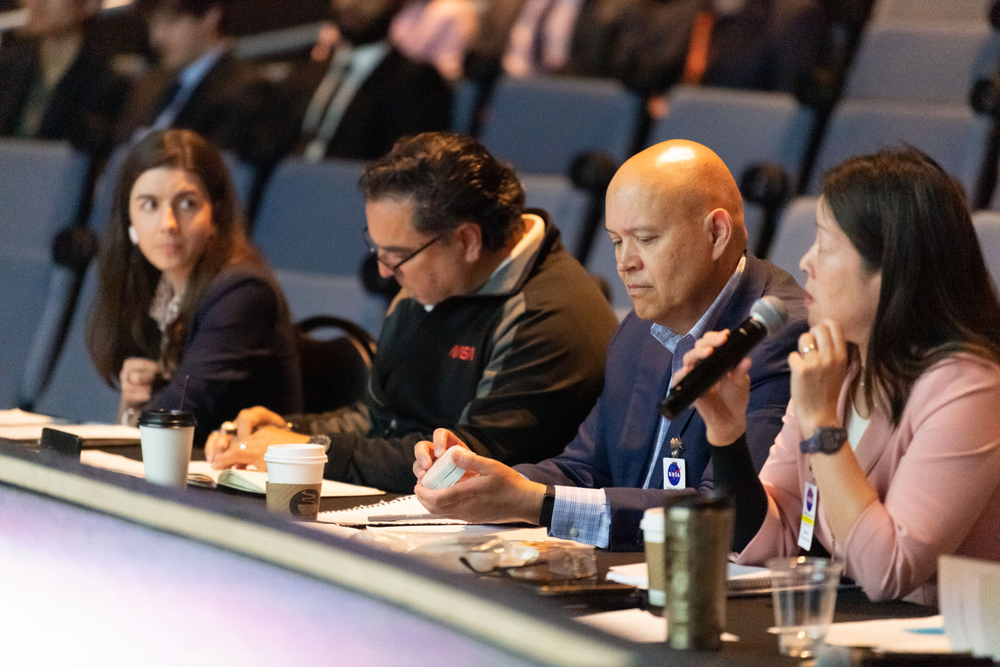 Four people sitting at a table looking through notes on the table. One person on the left is holding a microphone and speaking.