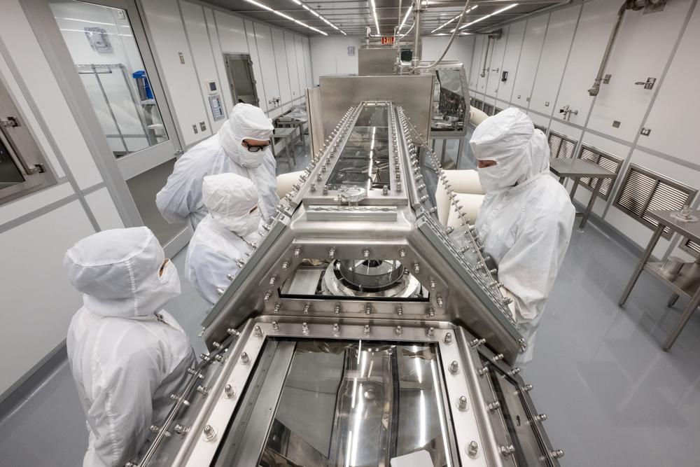 Four people dressed in white gowns inside of a laboratory looking inside of a container. 