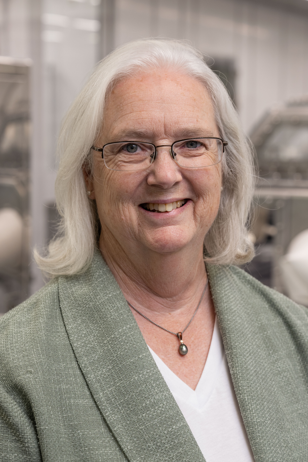 A person smiling inside of a facility. She is wearing a grey cardigan and white shirt.