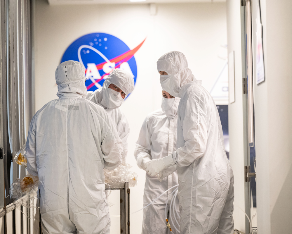 An image of four people in white gowns in a white room with a NASA meatball behind them.