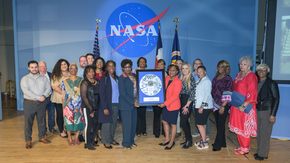 A group of people hold up an award with a blue background, NASA meatball, and three flags behind them. 