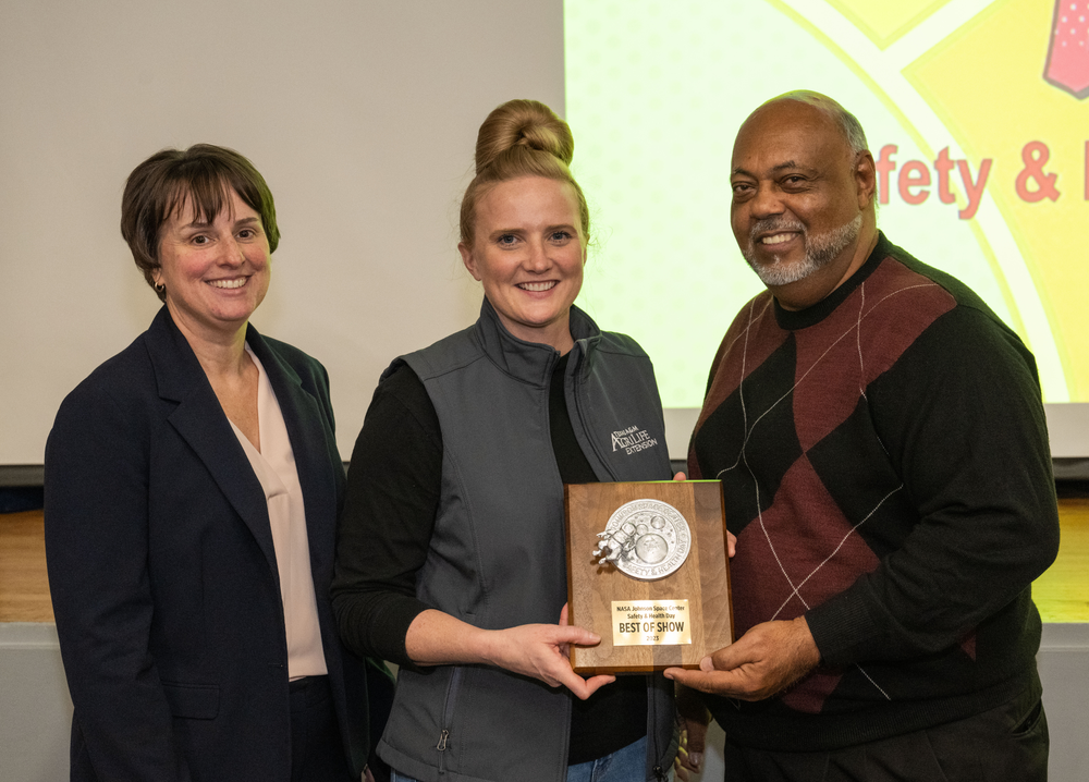 A woman wearing a black shirt and a grey vest accepts a wooden plaque from a man in a maroon-and-black sweater, with another woman in a black suit standing next to her.