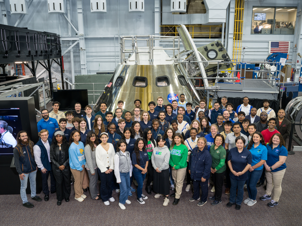 A group of people pose in a large facility with a spacecraft behind them.