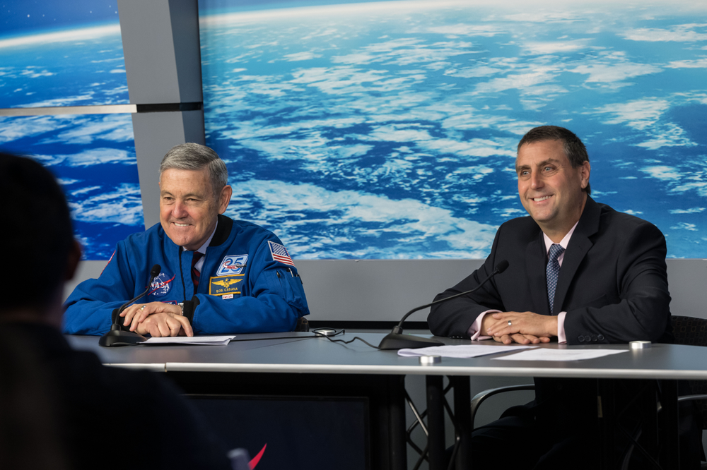 Two men, one wearing an astronaut jacket, sit at a table in front of a large image of the Earth taken from space.