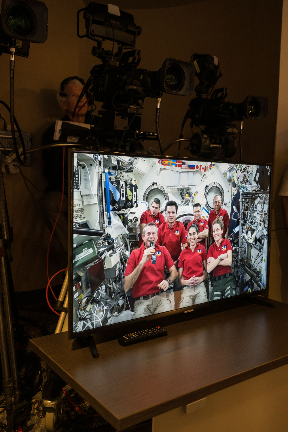 Seven astronauts wearing red polo shirts are shown on a television screen while floating onboard the International Space Station.