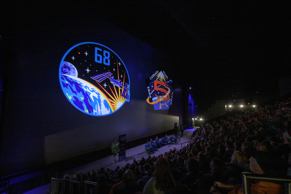 A dark-lit image of people sitting in an auditorium in front of a large screen. 
