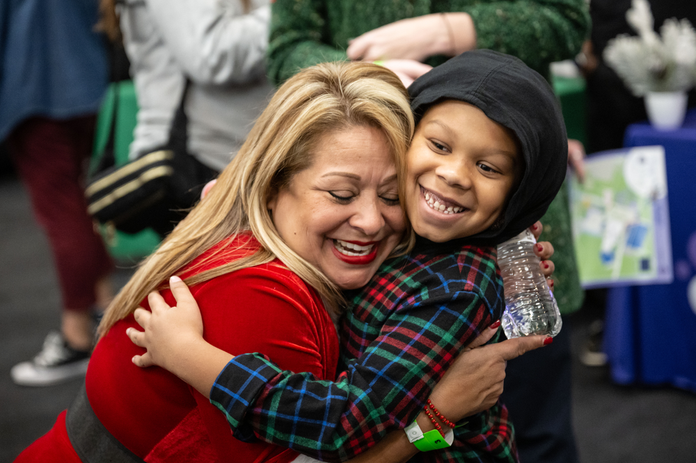 A person dressed as Santa in red hugs a child wearing a plaid shirt with a black hood. They both are smiling.