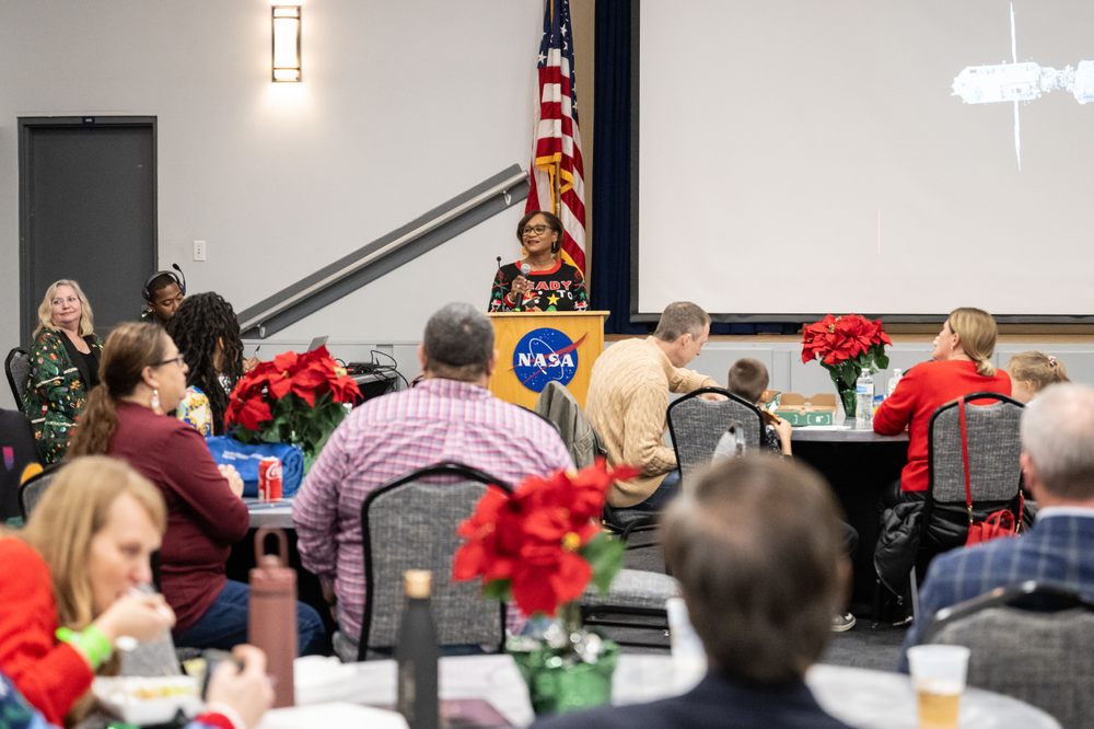 A person in a festive sweater stands at a podium to speak in front of a room of people sitting at tables. 