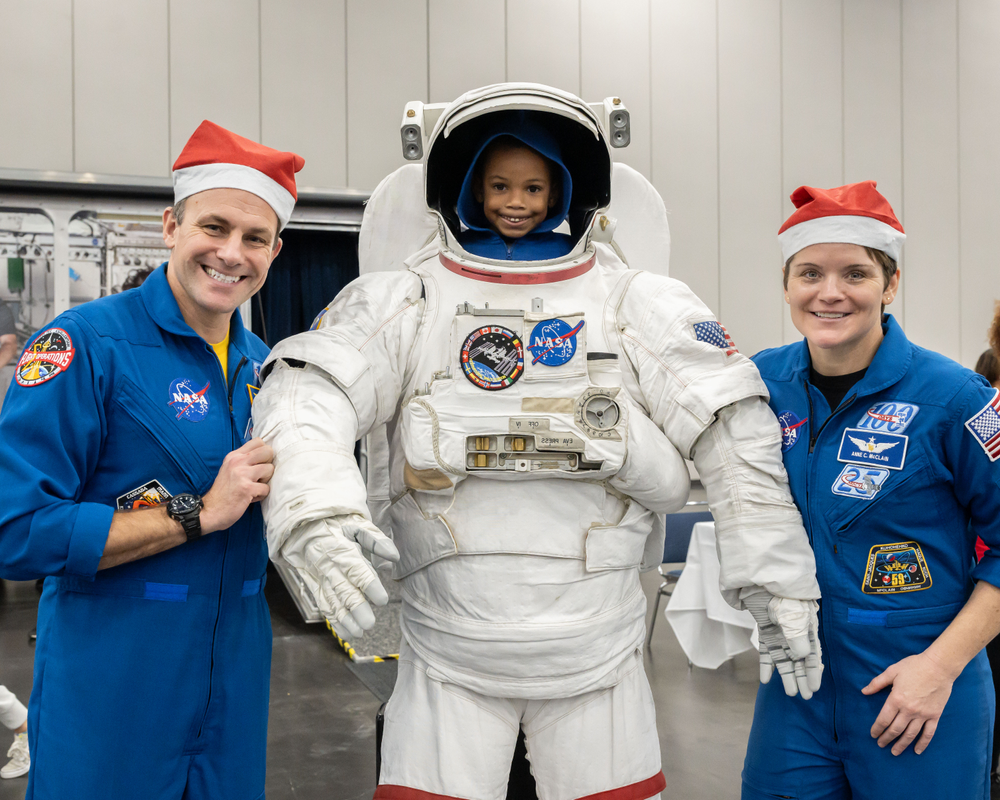 Two astronauts wearing blue flight suits and santa hats pose next to a young child who stands inside a spacesuit.