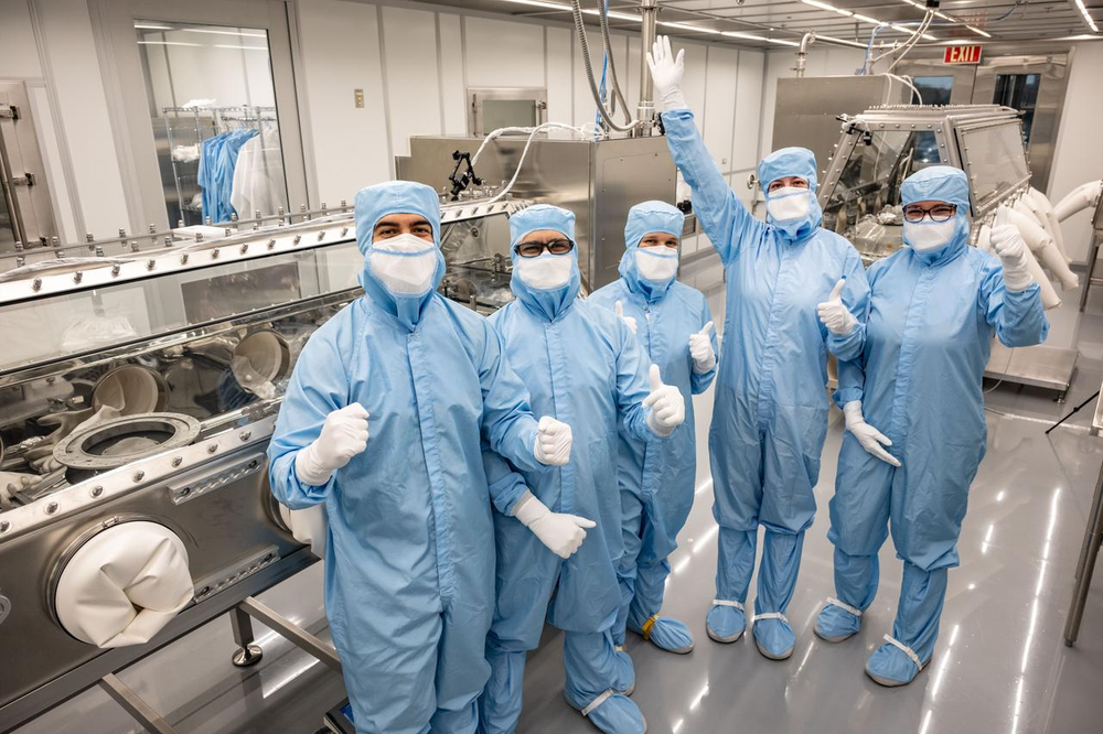 A group of scientists wearing blue protective suits and white face masks stands in a cleanroom at Johnson Space Center.