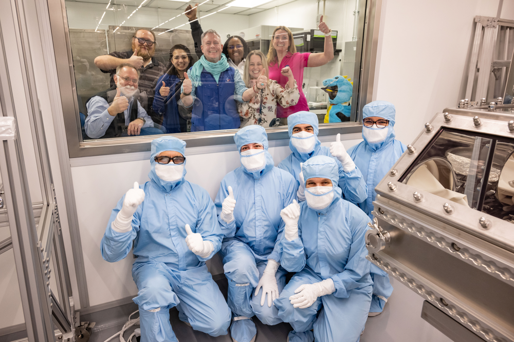 A group of five people dressed in blue gowns in a laboratory with seven people standing behind a glass behind them. 