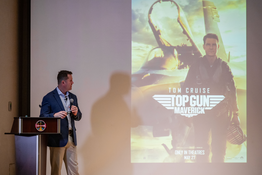 A person wearing a blue blazer and tan pants standing behind a podium in front of a screen. 