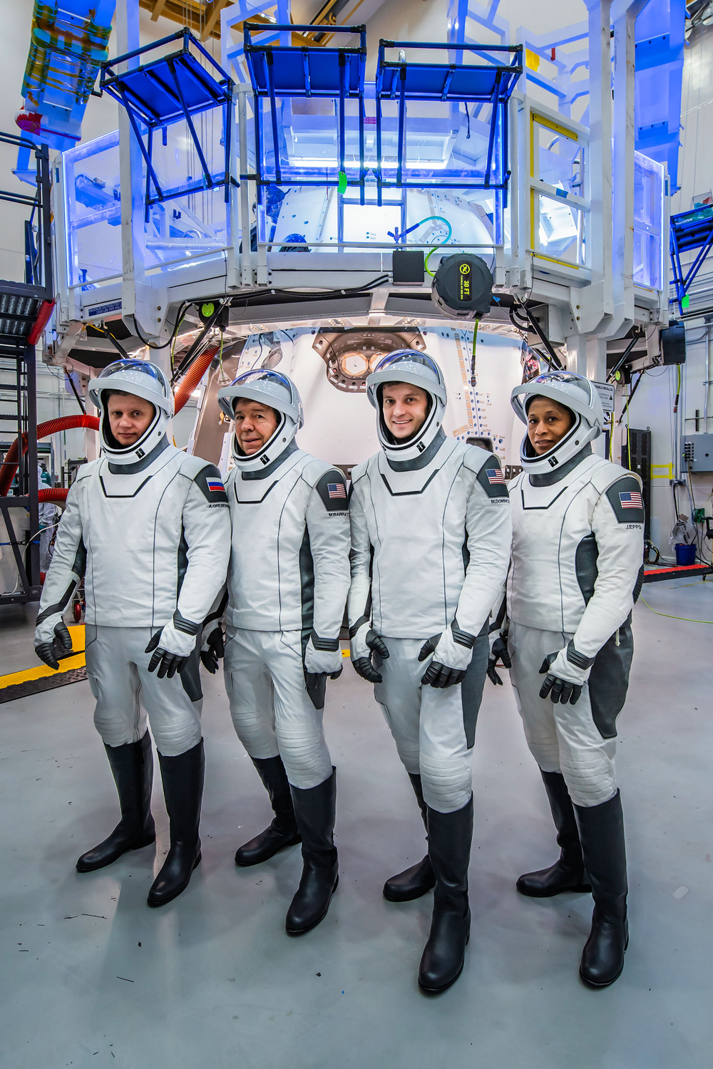 Four people dressed in white space suits pose in front of a large structure.