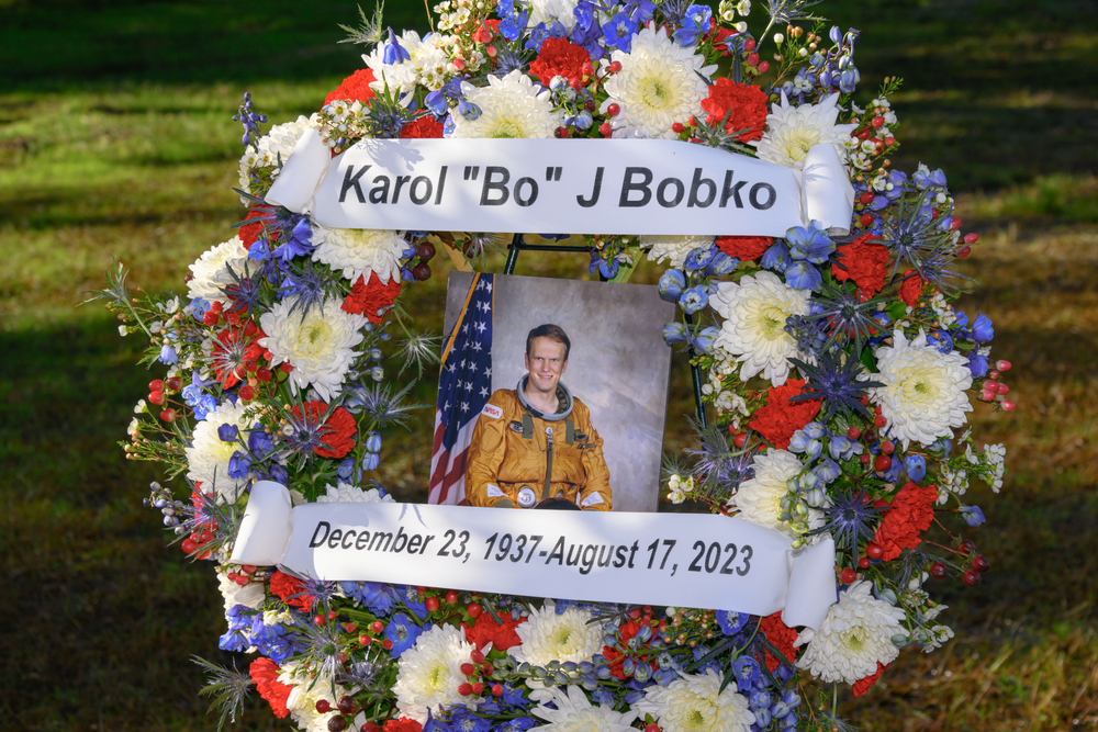 An image of a wreath containing flowers with an image of a person in an orange flight suit sitting next to an American flag in the middle. 