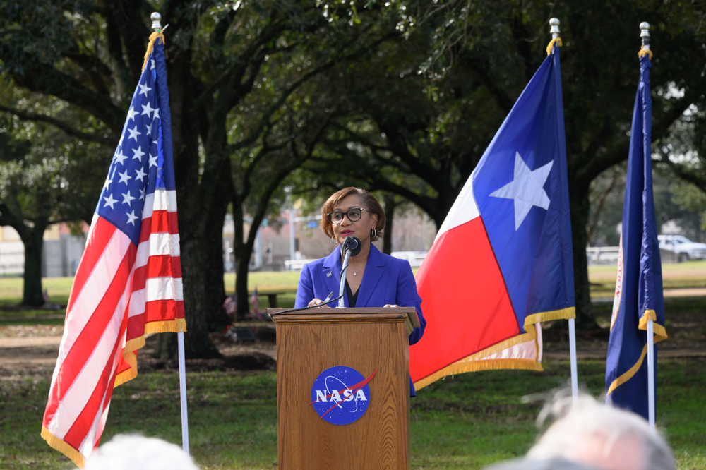 A person wearing a blue blazer standing in front of a podium and an American (left) and Texas (right) flag.