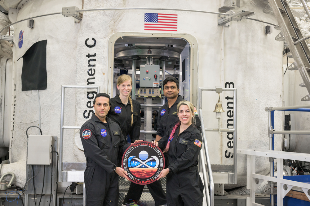 A group of four people in black spacesuits standing in front of a white habitat holding a crew patch. 