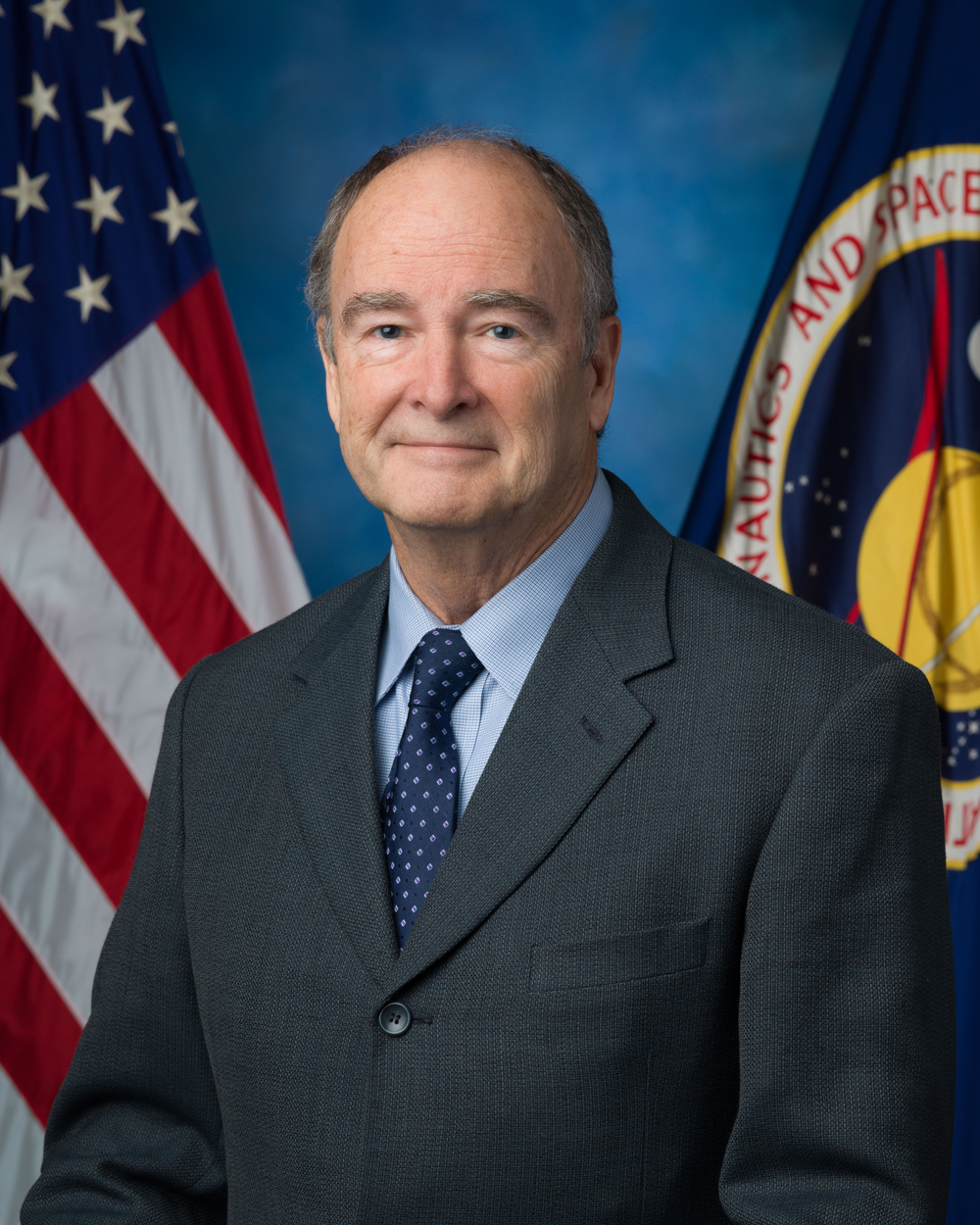 A person wearing a black suit and blue polka dot tie smiles in front of a blue background and a U.S. flag (left) and NASA flag (right).