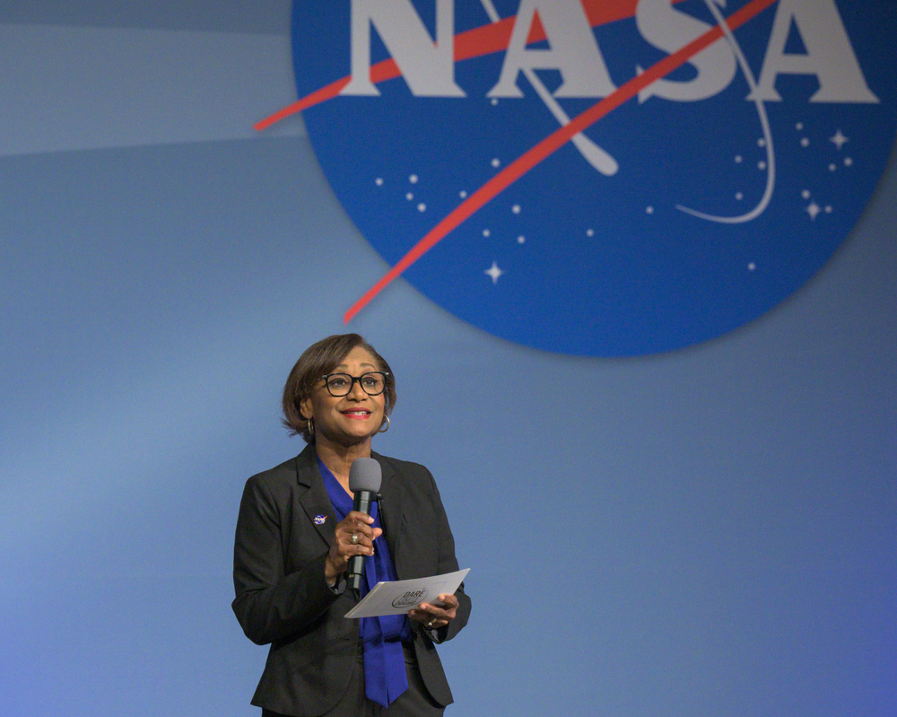 A woman wearing glasses, a black suit, and a blue shirt holds a microphone and note cards while speaking on a stage.