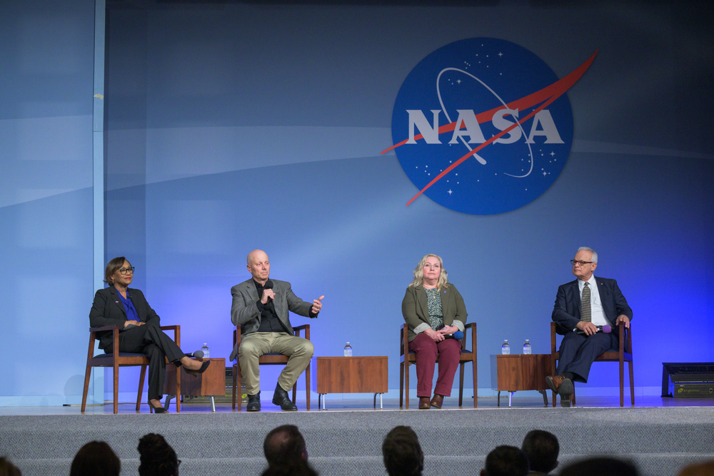 Two women and two men in professional attire sit on wooden chairs on a stage.