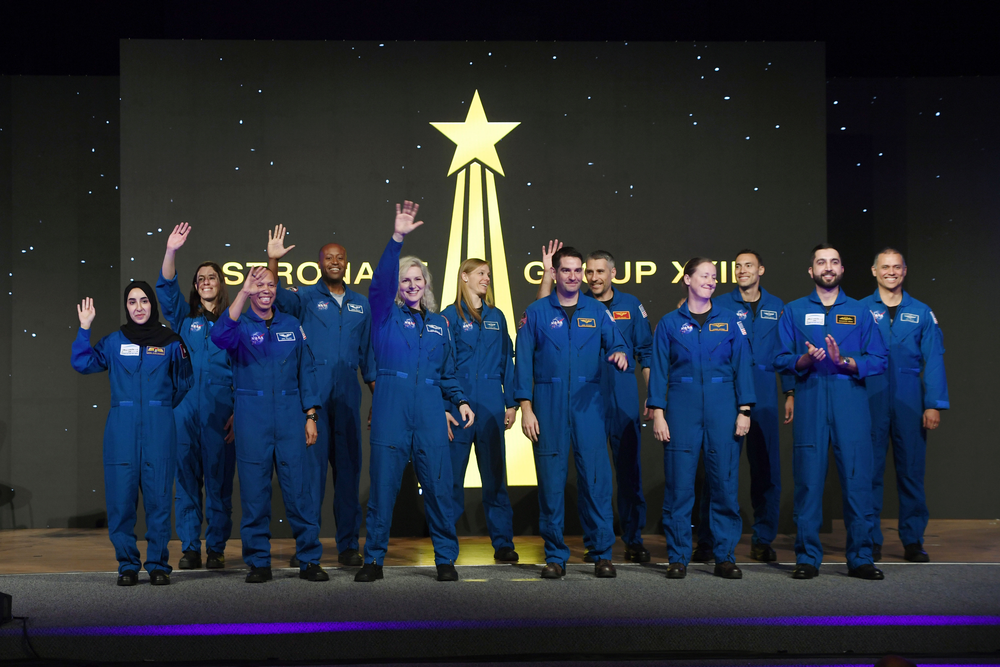 A group of people in blue spacesuits wave to the audience on a stage. The background is black with gold stars. 