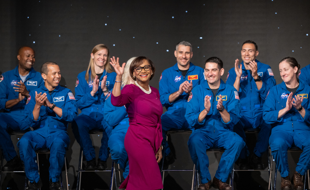 A group of people in blue spacesuits clap in the background as a person in a maroon dress waves and walks in front of them on a stage.