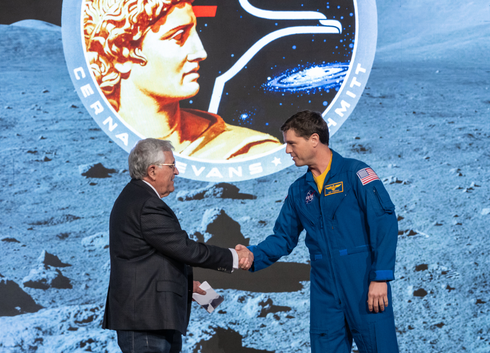 A person in a blue spacesuit, right, shakes hands with a person in a black suit, left, in front of a moon-themed background with a Greek and space-themed symbol.