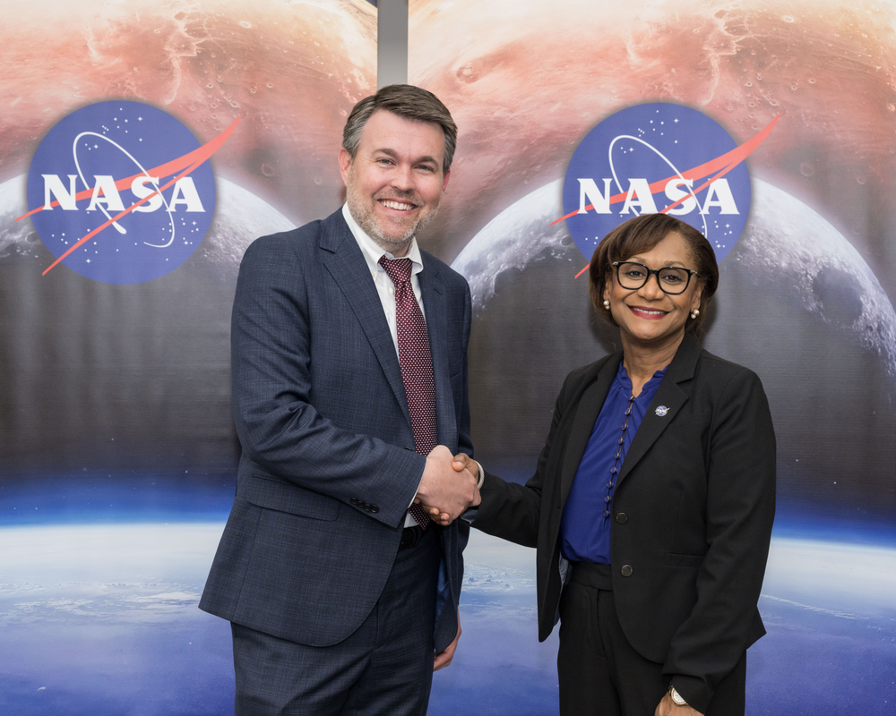 A white man with a beard and wearing a navy blue suit shakes hands with a Black woman wearing glasses and a black suit with a blue shirt in front of two NASA banners.