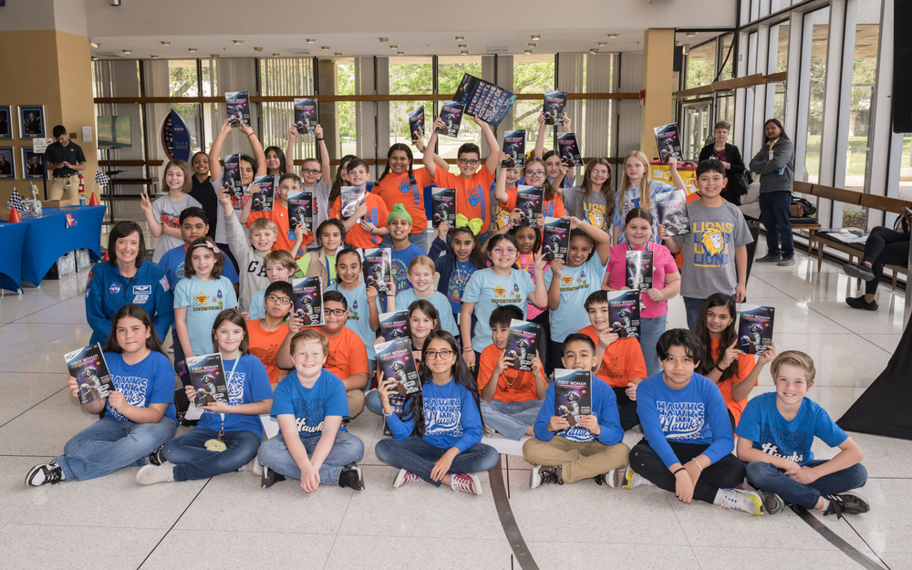 A group of children hold up posters and smile in a large, open room.