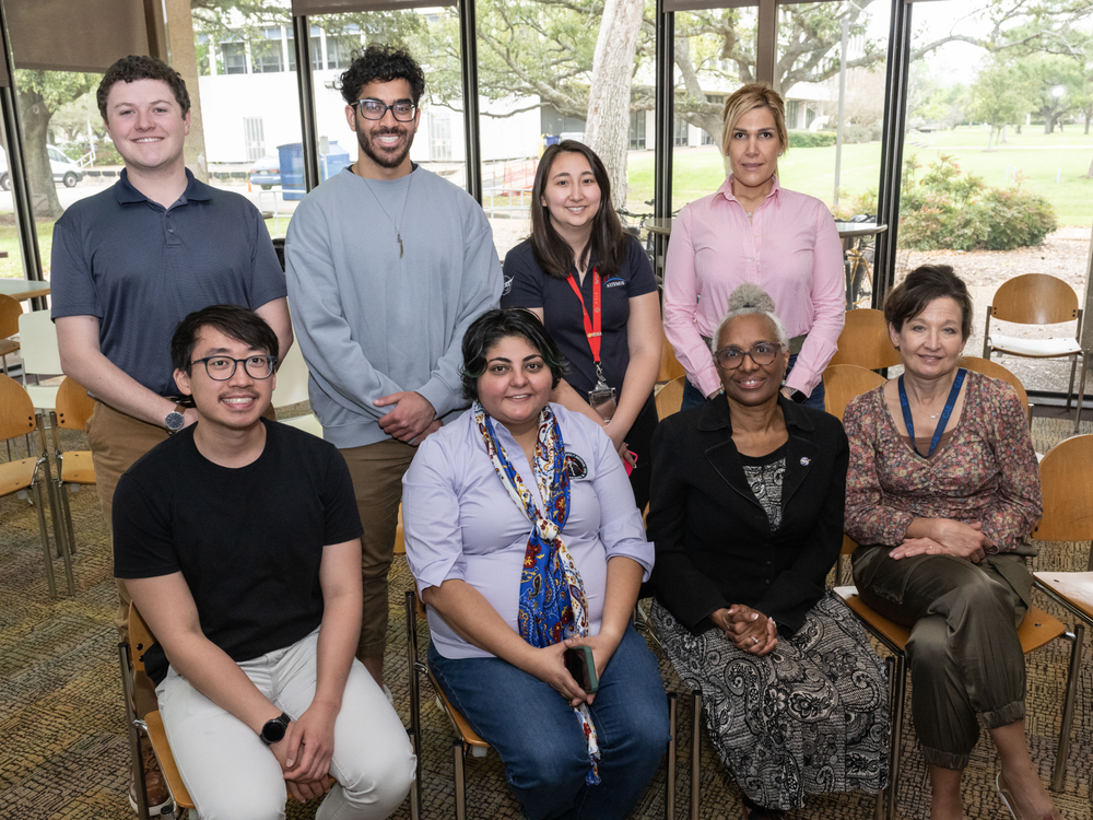 Eight people pose for a photo inside of a large cafeteria with windows behind them. 