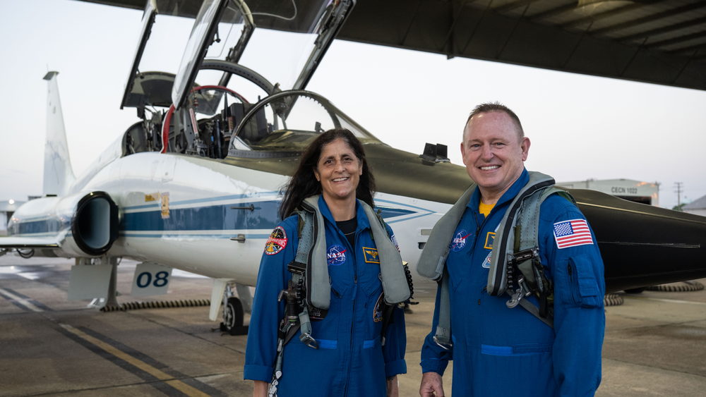 Two people dressed in blue flight suits stand outside in front of a jet.