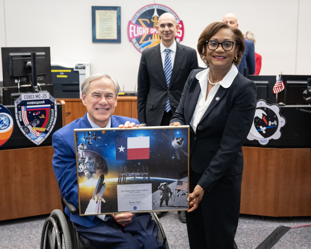 A white man with gray hair, wearing a blue suit, and sitting in a wheelchair receives a framed honorarium from a Black woman wearing a black suit and white button-down shirt.