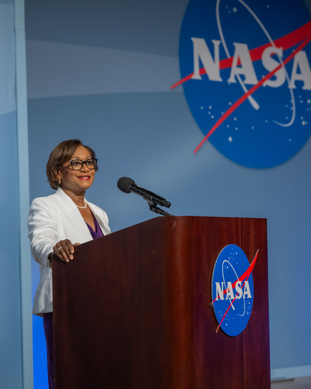 A Black woman with short hair and black-rimmed glasses and a white blazer stands behind a wooden podium.