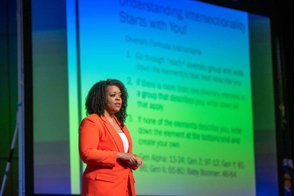 A black woman wearing an orange suit and white blouse stands in front of a large video screen delivering a presentation.